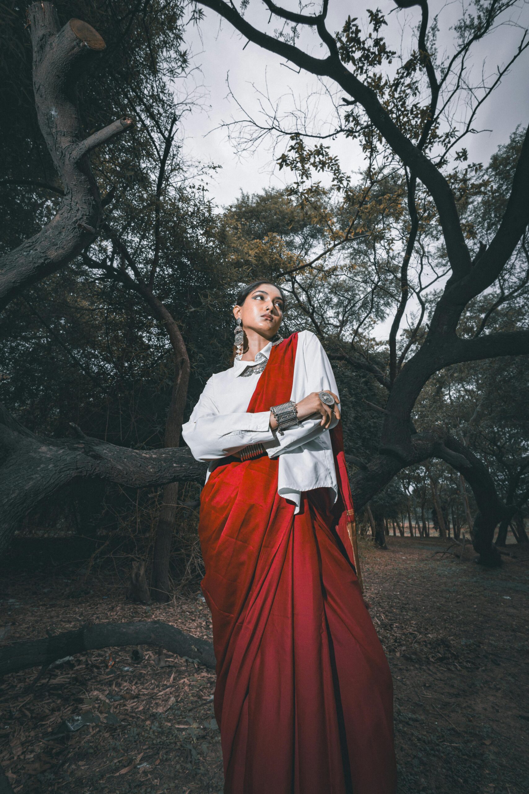 Portrait of a woman in a red saree and jewelry, standing in a forest with dramatic lighting.